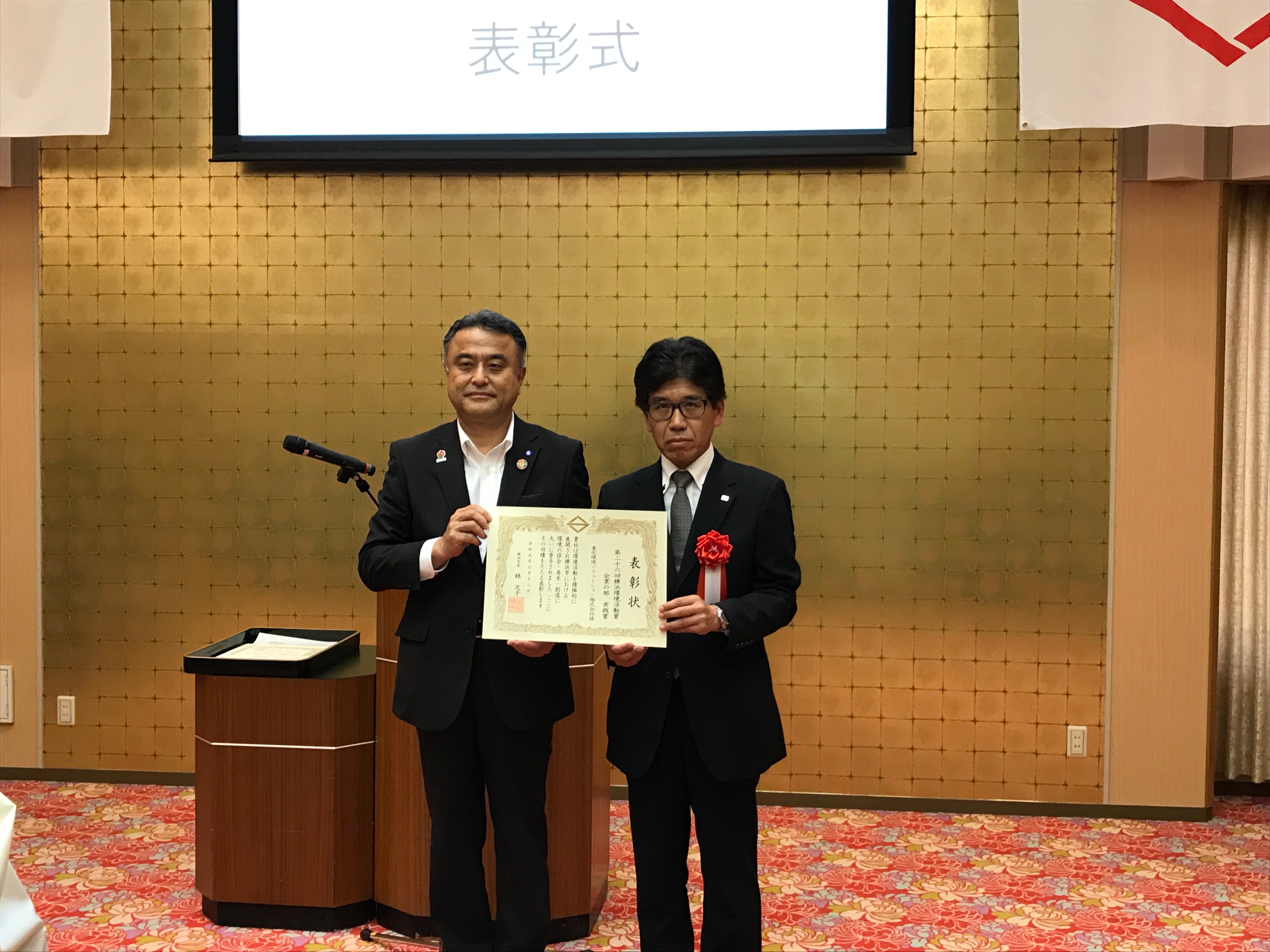 横浜市副市長 小林一美氏(左）より表彰状を授与される当社経営企画部長 鈴木茂明